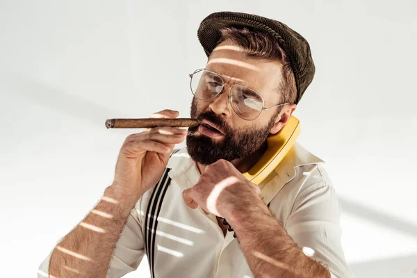 Красивый мужчина в очках и кепке разговаривает по винтажному телефону и курит сигары — стоковое фото