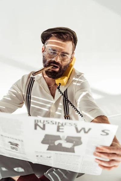 Handsome bearded man in glasses talking on phone, smoking cigar and reading business newspaper — Stock Photo