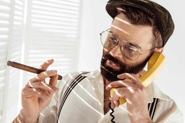 Homme barbu dans des lunettes et casquette parler au téléphone et tenant cigare — Photo de stock