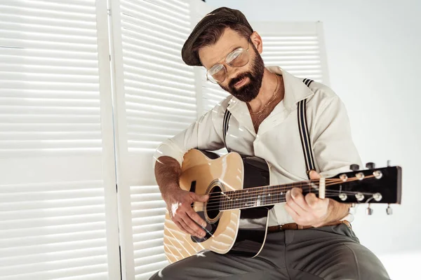 Hombre barbudo guapo en gorra y gafas sentado y tocando la guitarra cerca del divisor de habitación blanca - foto de stock