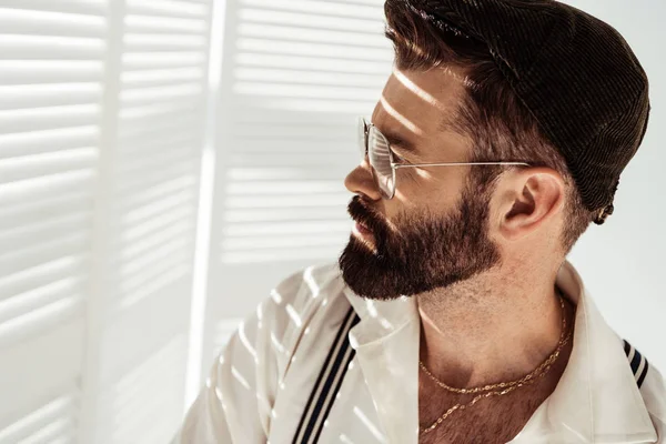 Bel homme barbu dans des lunettes et un chapeau près de diviseur de pièce blanche — Photo de stock