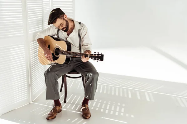 Bonito barbudo homem no cap sentado na cadeira e tocando guitarra perto de branco sala divisor — Fotografia de Stock