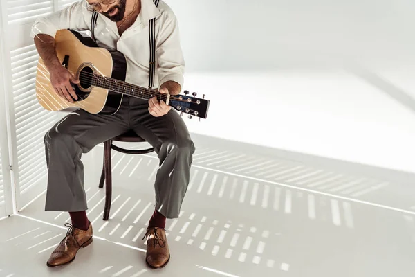 Бородатий чоловік в окулярах сидить на стільці і грає на акустичній гітарі біля роздільника білої кімнати — стокове фото