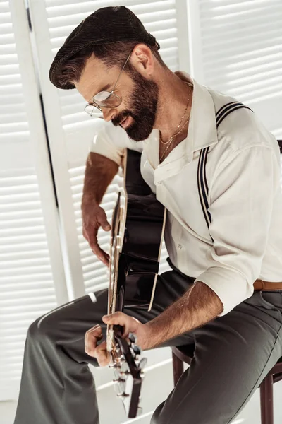 Bonito barbudo homem sentado na cadeira e tocando guitarra acústica perto de tela dobrável no quarto — Fotografia de Stock