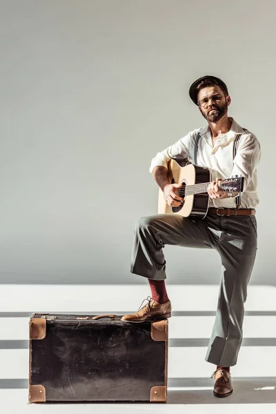 Бородатый мужчина в кепке рядом с винтажным чемоданом играет на акустической гитаре и смотрит на камеру на сером фоне — стоковое фото