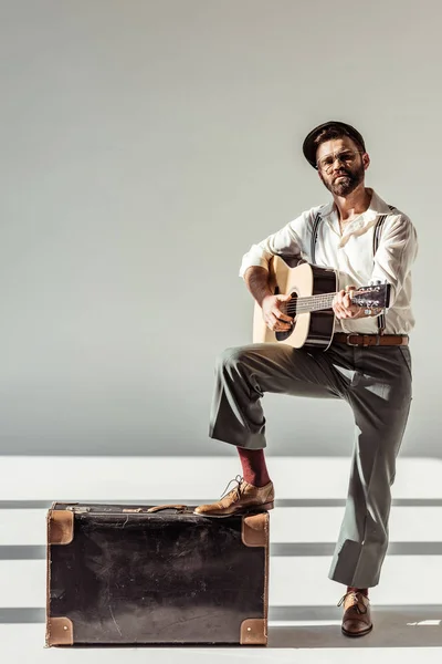 Barbudo hombre con gorra cerca de la maleta vintage tocando la guitarra acústica y mirando a la cámara sobre fondo gris - foto de stock