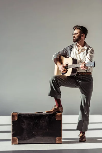 Bärtiger Musiker mit Mütze in der Nähe eines Vintage-Koffers, der Akustikgitarre auf grauem Hintergrund spielt — Stockfoto