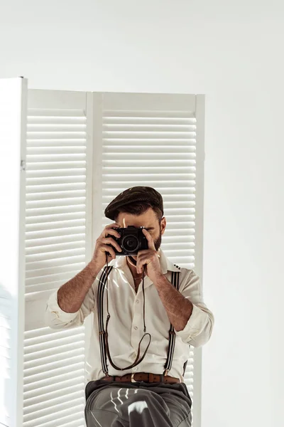 Hombre barbudo sentado cerca del divisor de habitación blanca tomando fotos con cámara de película vintage - foto de stock