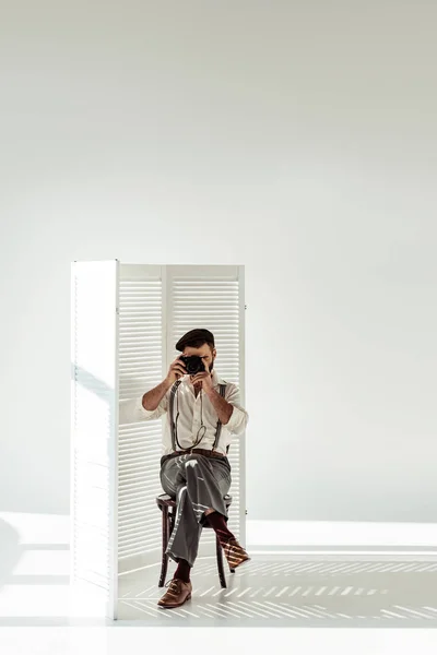 Bearded man sitting on chair near room divider and taking pictures with vintage film camera — Stock Photo