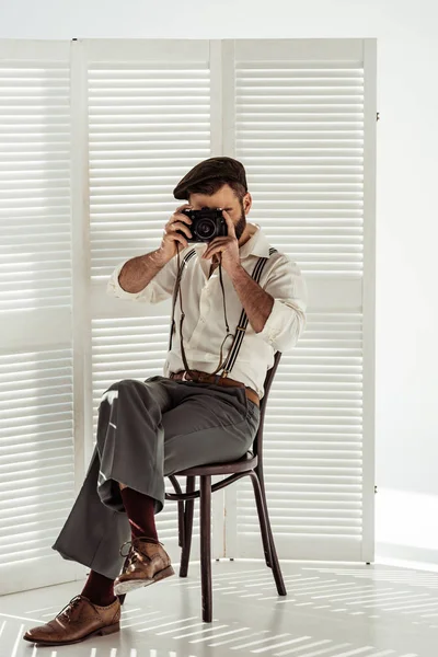 Homem barbudo sentado na cadeira e tirando fotos com câmera de filme vintage — Fotografia de Stock