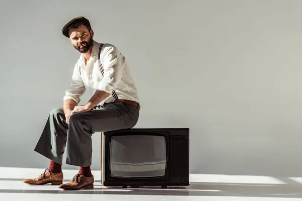 Hombre barbudo guapo sentado en la televisión vintage y mirando a la cámara en el fondo gris - foto de stock