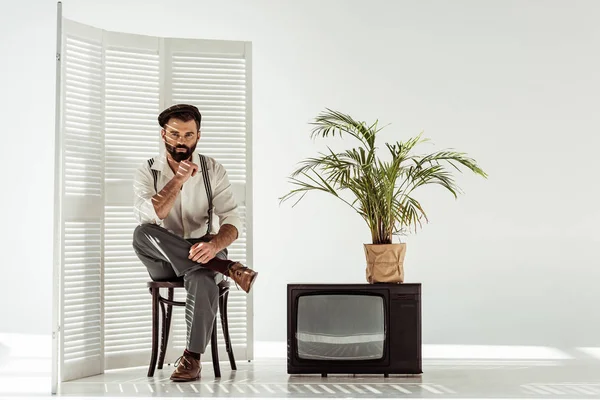 Bonito barbudo homem sentado na cadeira perto de tela dobrável e tv no branco quarto — Fotografia de Stock