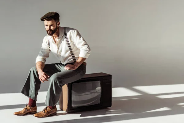 Guapo hombre barbudo con estilo en la tapa sentado en la televisión vintage sobre fondo gris - foto de stock