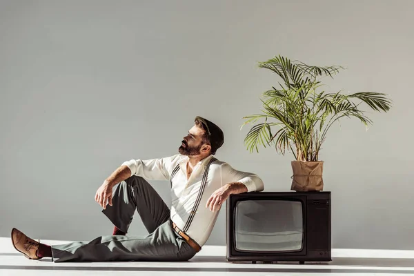 Hombre barbudo guapo sentado en el suelo cerca de la televisión vintage con planta en maceta - foto de stock