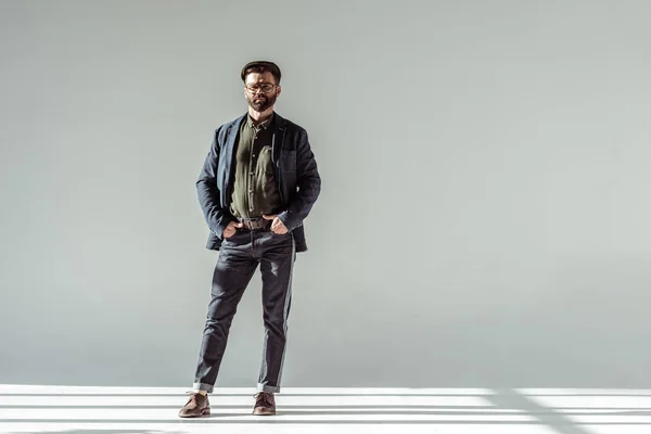 Schöner Mann mit Mütze, die Hände in den Taschen stehend und in die Kamera blickend auf grauem Hintergrund — Stockfoto