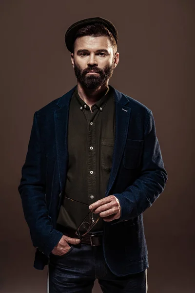 Handsome bearded man with hand in pocket holding glasses and looking at camera isolated on brown — Stock Photo