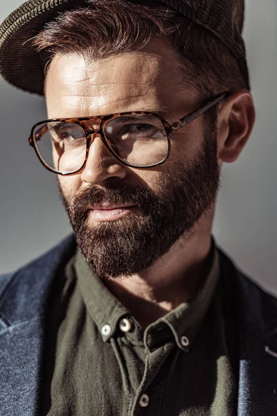 Vista de cerca del hombre barbudo guapo en gafas y gorra mirando a la cámara aislada en gris - foto de stock