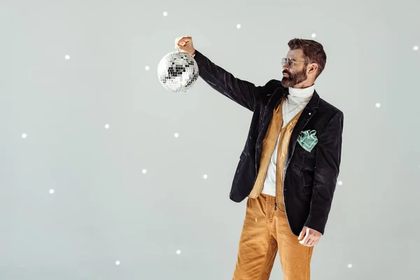 Красивый бородатый мужчина смотрит на диско-шар в руке на сером фоне — стоковое фото