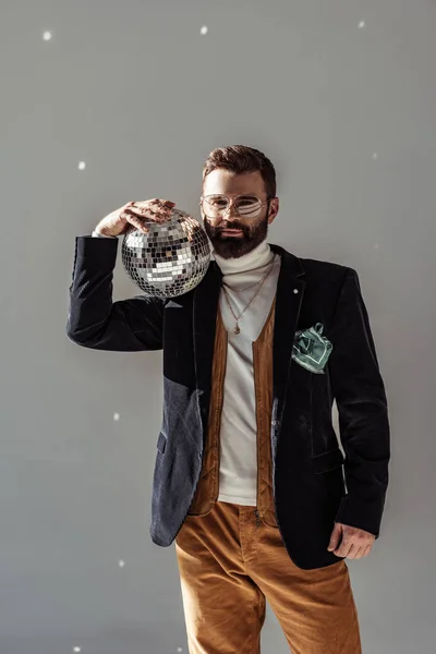 Handsome bearded man in glasses holding disco ball on shoulder and looking at camera on grey background — Stock Photo