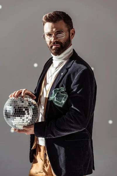 Schöner Mann mit Brille, Discokugel in der Hand und Blick in die Kamera auf grauem Hintergrund — Stockfoto