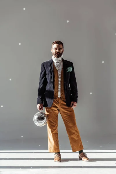 Bearded man standing, holding disco ball and looking at camera on grey background — Stock Photo