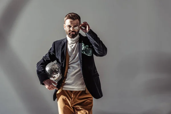 Bearded man touching glasses, holding mosaic disco ball and looking at camera on grey background — Stock Photo