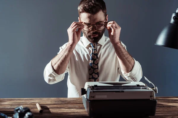 Bärtiger Journalist, der mit Schreibmaschine am Tisch sitzt, Brille anfasst und auf grauen Hintergrund in die Kamera blickt — Stockfoto