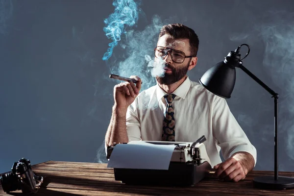 Nachdenklicher bärtiger Journalist sitzt mit Schreibmaschine am Tisch und raucht vor grauem Hintergrund — Stockfoto