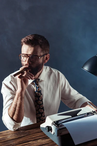 Nachdenklicher bärtiger Journalist sitzt mit Retro-Schreibmaschine am Tisch und raucht vor grauem Hintergrund — Stockfoto