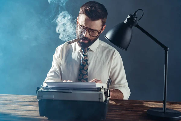 Красивый журналист в очках сидит за столом, печатает на винтажной пишущей машинке и курит на сером фоне — стоковое фото
