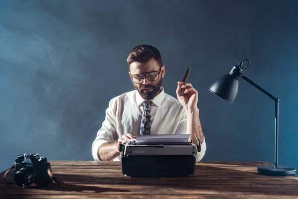 Красивый журналист в очках сидит за столом с ретро пишущей машинкой и курит на сером фоне — стоковое фото