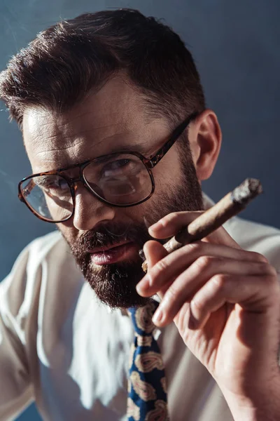 Handsome man in glasses holding cigar and looking at camera on grey background — Stock Photo