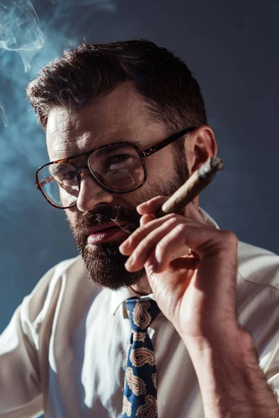 Hombre barbudo serio en gafas sosteniendo cigarro y mirando a la cámara sobre fondo gris - foto de stock