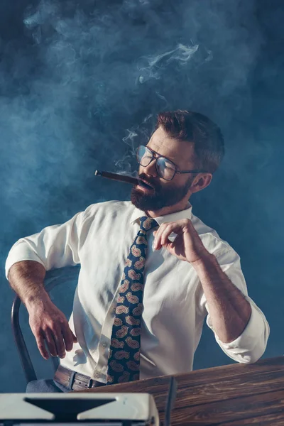 Pensativo periodista barbudo en gafas sentado a la mesa con máquina de escribir vintage y fumar sobre fondo gris - foto de stock