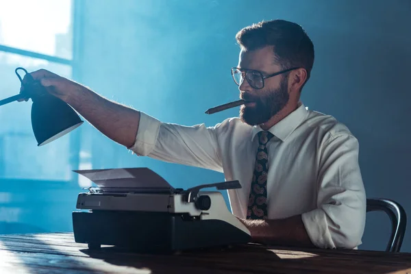 Бородатый журналист в очках сидит за столом со старинной пишущей машинкой, курит и трогает лампу возле окна — стоковое фото