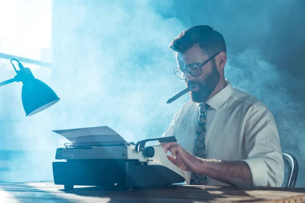 Periodista barbudo en gafas sentado en la mesa, escribiendo en la máquina de escribir vintage y fumar cerca de la ventana - foto de stock