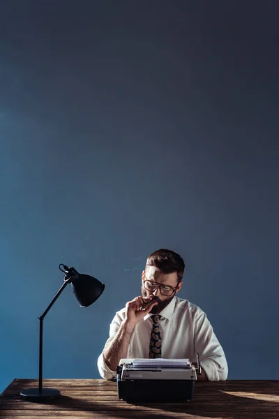 Schöner Journalist sitzt mit Lampe und Retro-Schreibmaschine am Tisch und raucht vor grauem Hintergrund — Stockfoto