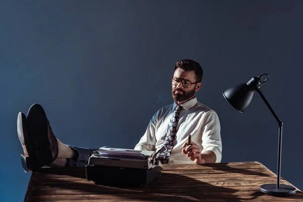 Bärtiger Journalist stellt Füße auf den Tisch, blickt auf alte Schreibmaschine und hält Zigarre auf grauem Hintergrund — Stockfoto