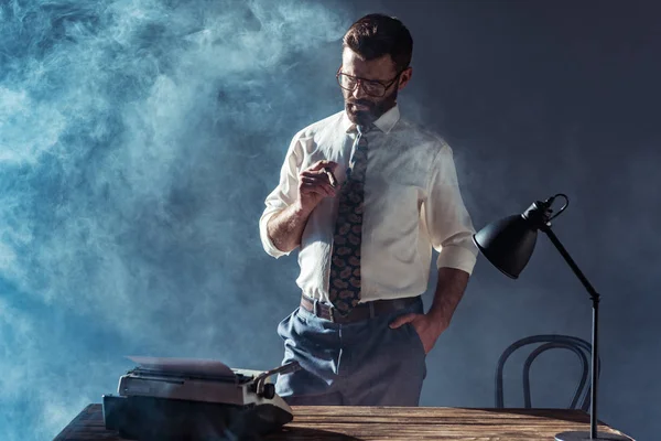 Красивий бородатий чоловік в окулярах тримає сигару і дивиться на друкарську машинку — стокове фото
