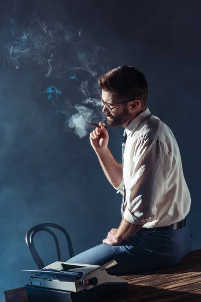 Schöner bärtiger Mann mit Brille sitzt auf dem Tisch, während er Zigarre raucht — Stockfoto