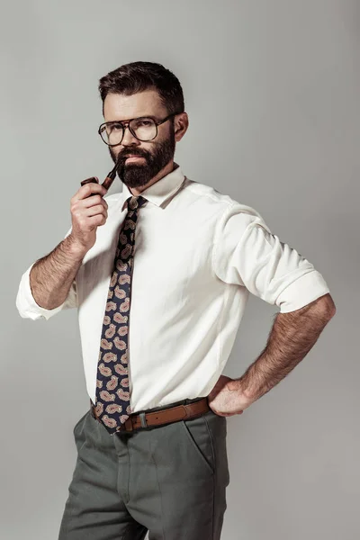 Красивий бородатий чоловік в окулярах і сорочці курить трубу і дивиться на камеру ізольовано на сірому — стокове фото