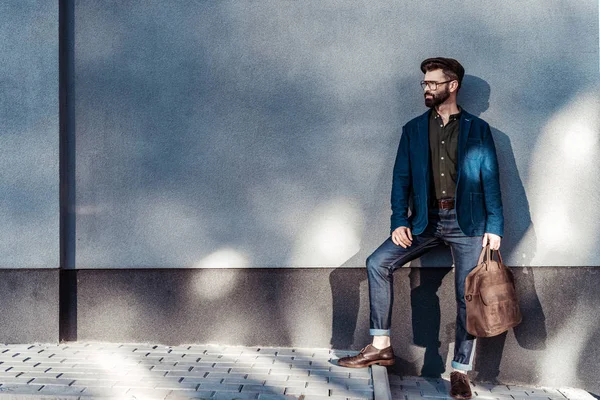 Handsome bearded man in glasses and cap holding bag near wall — Stock Photo