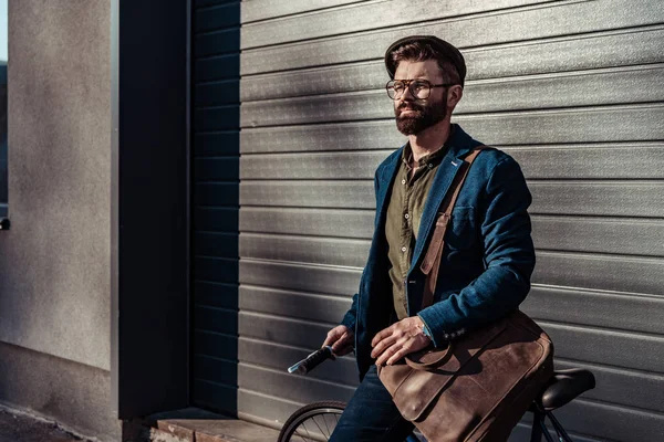 Homem barbudo bonito em óculos e boné segurando bicicleta e sorrindo — Fotografia de Stock