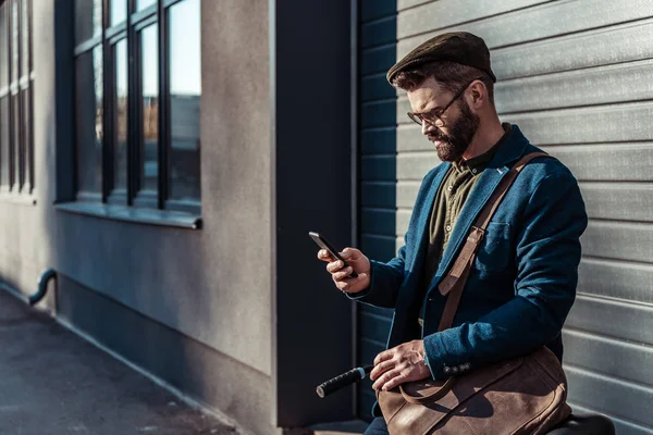 Handsome bearded man in glasses and cap holding bicycle and looking at smartphone — Stock Photo