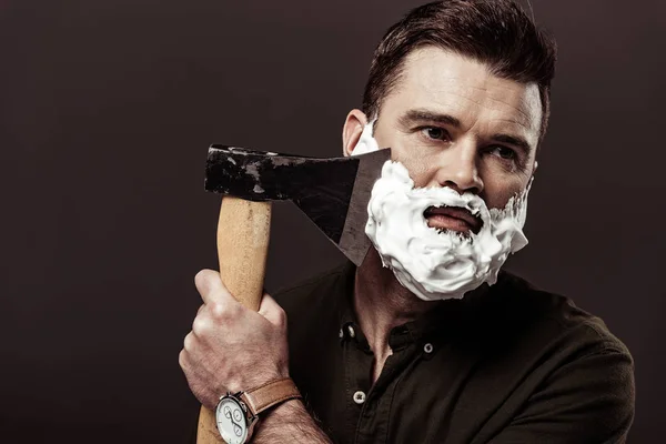 Handsome bearded man in brown shirt shaving with ax isolated on brown — Stock Photo