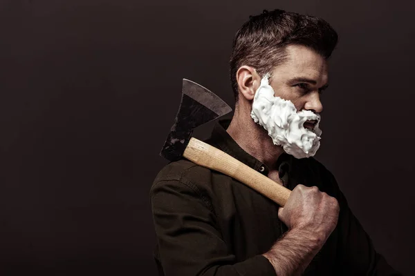 Красивий бородатий чоловік з піною для гоління на обличчі тримає сокиру ізольовано на коричневому — стокове фото