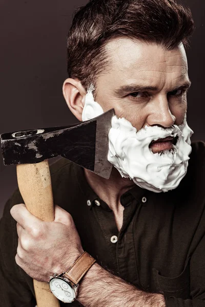 Hombre guapo serio con espuma en la cara barba de afeitar con hacha aislada en marrón - foto de stock
