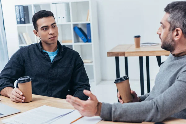 Professional businessmen holding paper cups and talking while sitting at workplace — Stock Photo