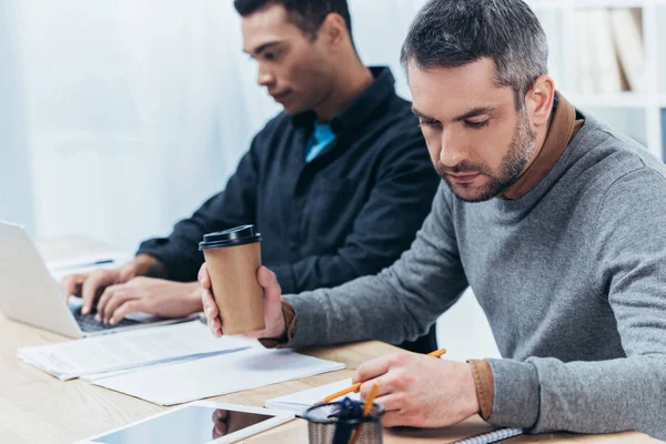 Homme d'affaires sérieux tenant café pour aller et prendre des notes tandis que le jeune collègue en utilisant ordinateur portable derrière — Photo de stock