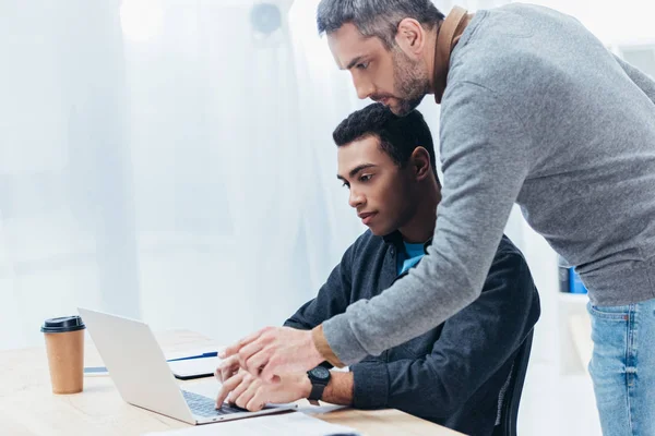 Mentor barbudo ayudar a joven colega que trabaja con el ordenador portátil en la oficina - foto de stock